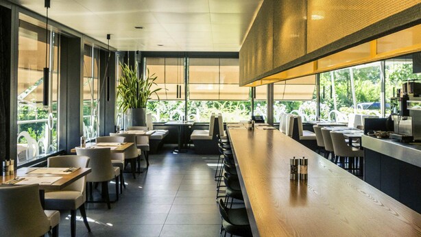 Un restaurante moderno con una larga barra de madera, varias mesas y sillas. Amplias ventanas ofrecen vistas al exterior.