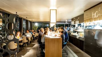 A modern restaurant with a long communal table, individual seating, and a kitchen area visible to the right.