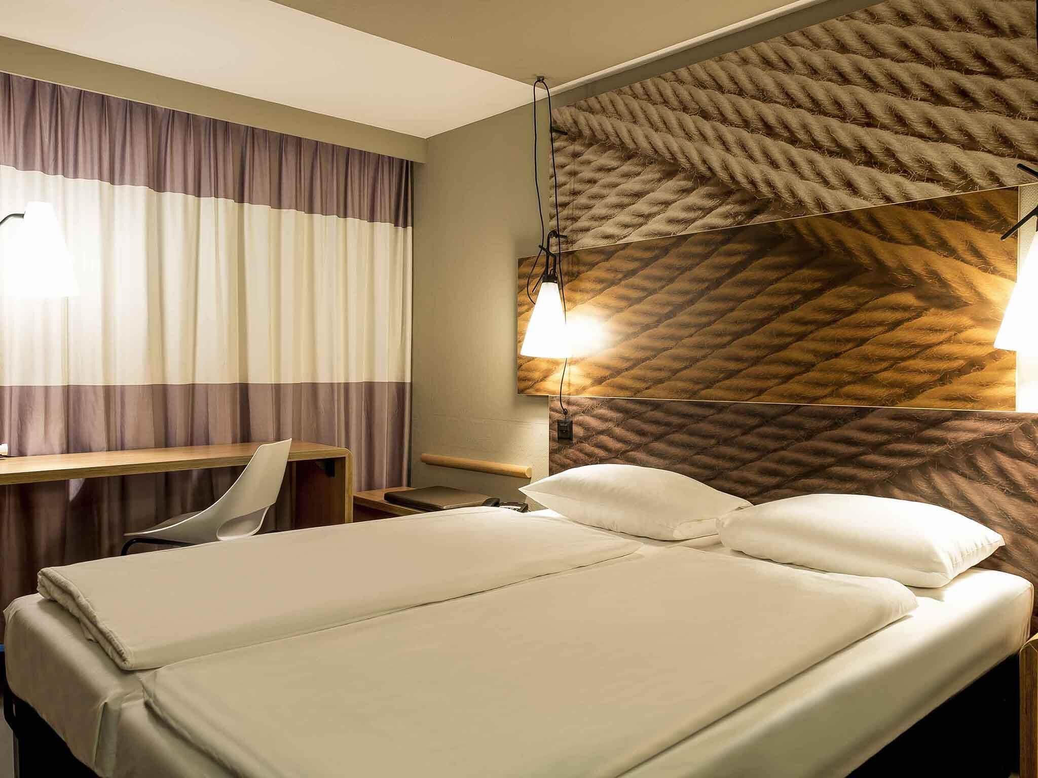 A hotel room with a bed, a desk, a chair, and a wall-mounted light fixture.