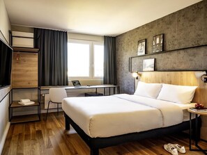 A hotel room with a bed, desk, chair, and a view of the outside. - ibis Zurich Adliswil (Adliswil)