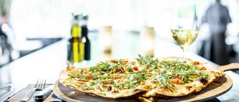 A pizza on a wooden board with a fork and knife beside it, and a glass of white wine in the background.