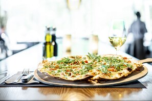 Una pizza sobre una tabla de madera, con un tenedor y un cuchillo a un lado, y una copa de vino blanco al fondo.