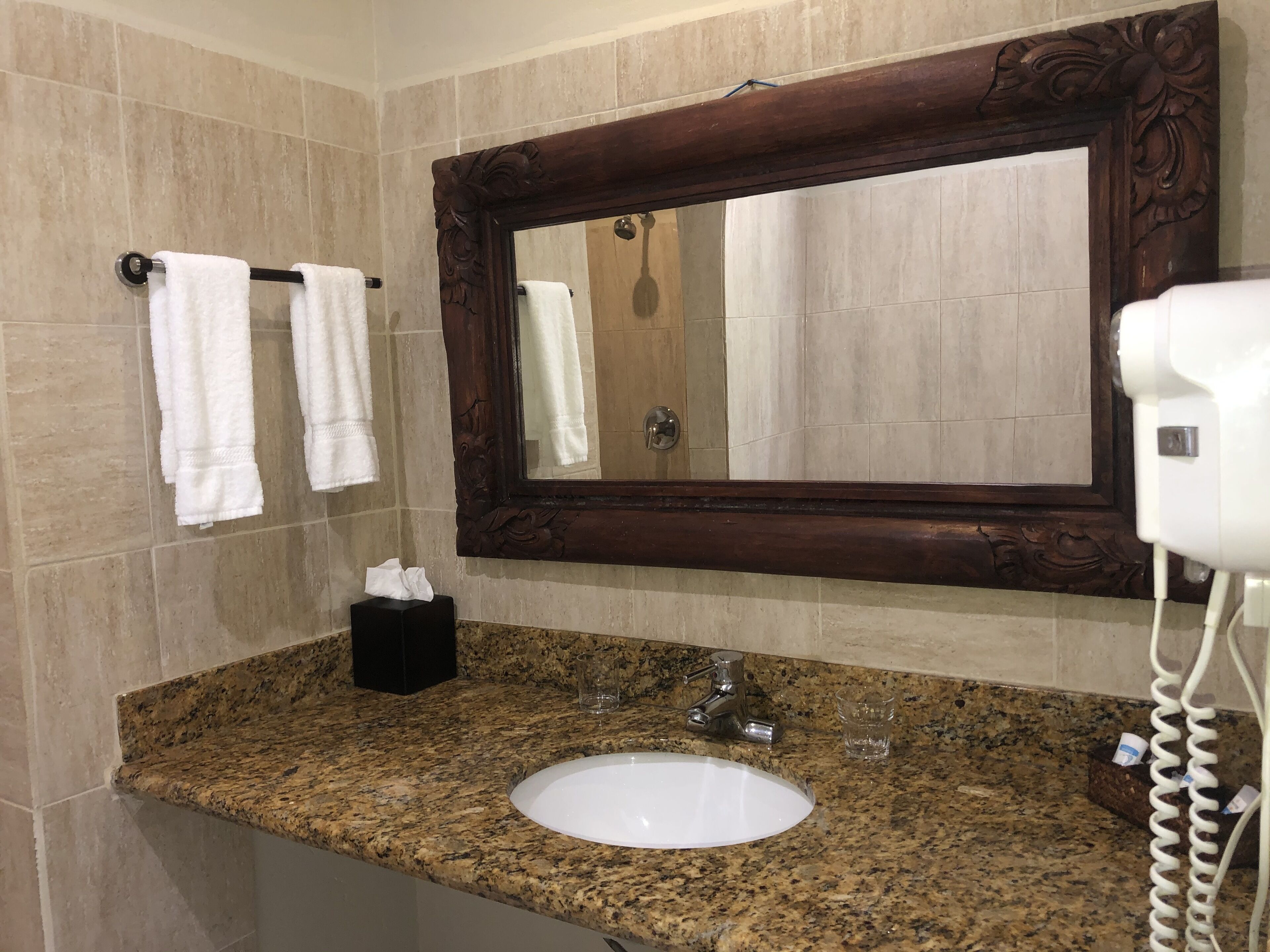 A bathroom with a granite countertop, a white sink, a mirror with an ornate frame, two white towels, a tissue box, and a hairdryer.