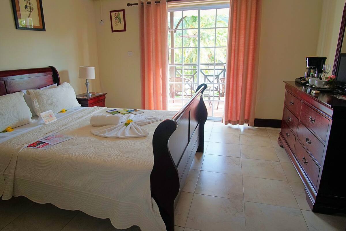 A hotel room with a neatly made bed, a wooden dresser, a coffee maker, and a view of the outdoors through a window with curtains.