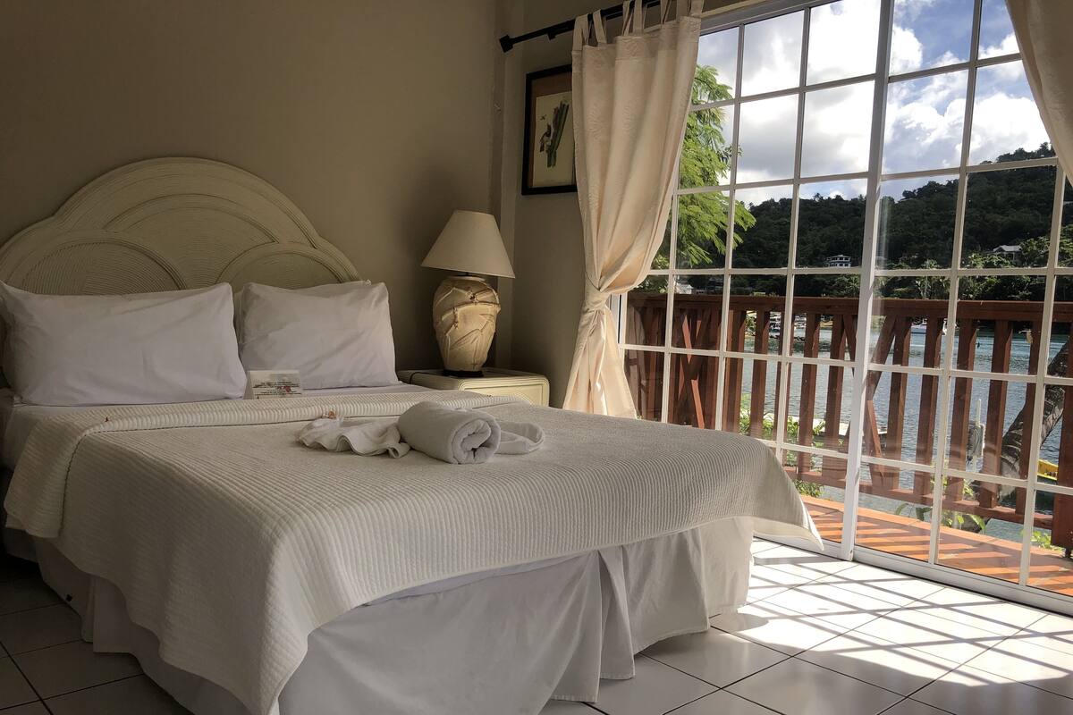 A neatly made bed with white linens and a rolled towel, a bedside lamp, and a window with curtains.