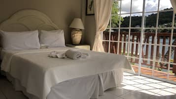 Una cama bien tendida con sábanas blancas y una toalla enrollada, una lámpara de noche y una ventana con cortinas.