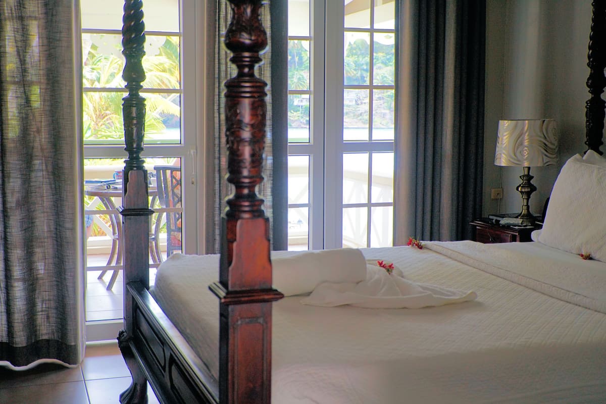 A four-poster bed with a white bedspread, a nightstand with a lamp, and a view of palm trees through the window.