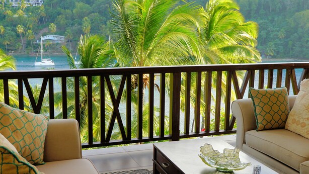A balcony with a view of a beach and palm trees. There is a sofa and a coffee table with a remote control on it.