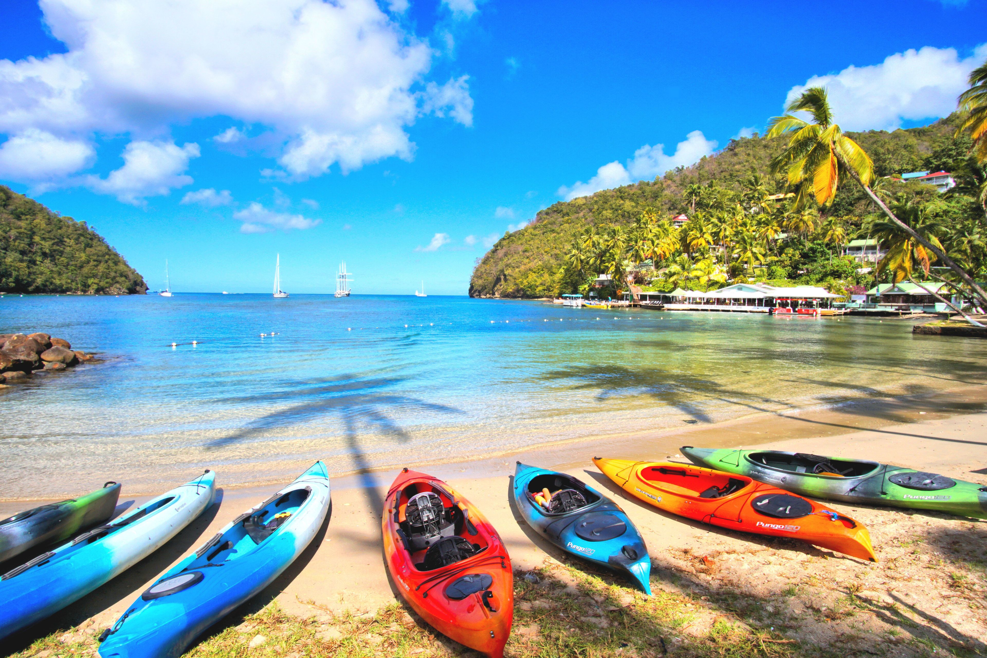 A beach with kayaks lined up on the sand, a clear blue sea, and a distant sailboat.