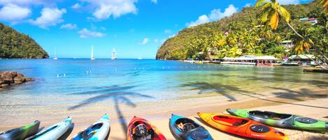 A beach with kayaks lined up on the sand, a clear blue sea, and a distant sailboat.