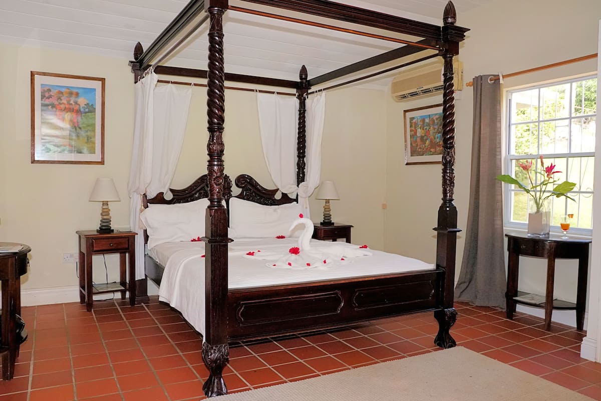 A four-poster bed with white bedding, a small bedside table with a lamp, and a window with curtains.