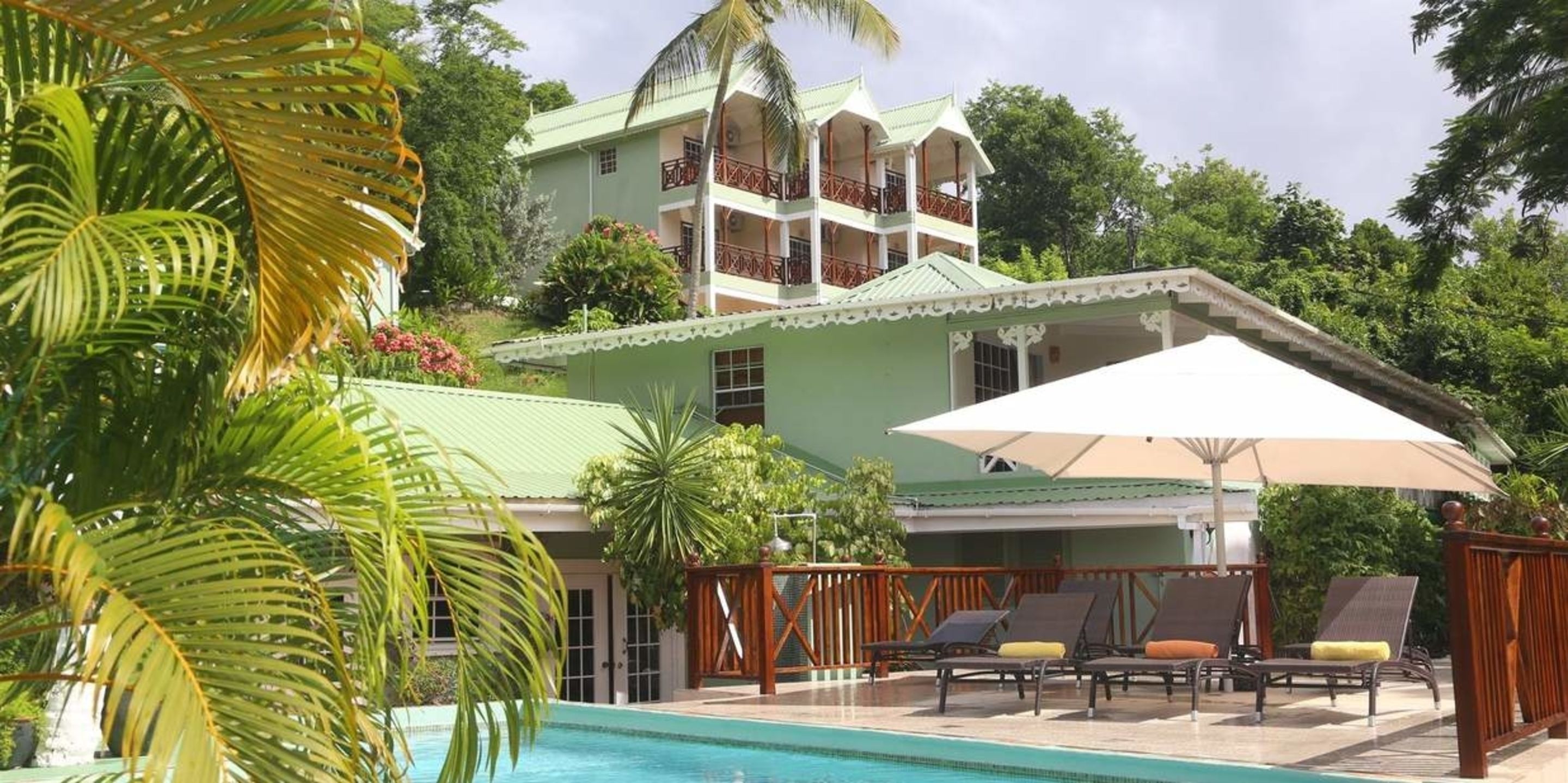 A pool area with lounge chairs and umbrellas, a multi-story building with balconies, and lush greenery.