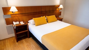 A hotel room with a wooden headboard, a white bedspread, two yellow pillows, a bedside table with a lamp, a phone, and a notepad. - Bristol (Buenos Aires)