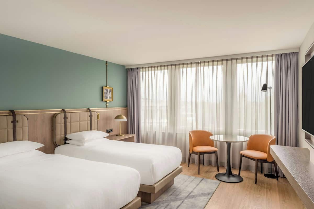 Hotellihuone, jossa on kaksi sänkyä, pieni pöytä tuoleineen, televisio ja ikkuna, jossa on verhot.