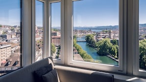 Una habitación con amplios ventanales que ofrecen vistas a un paisaje urbano, incluyendo un río y montañas.