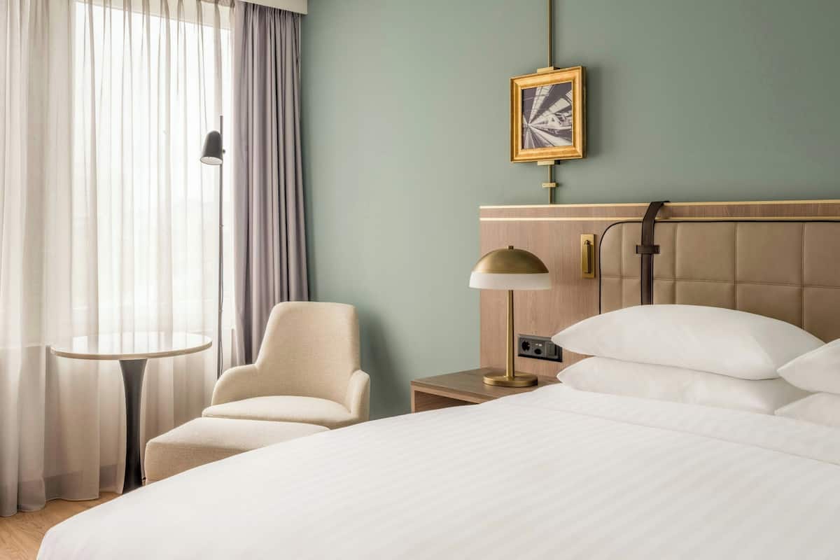 Hotellihuone, jossa on sänky, tuoli, pieni pöytä, lamppu ja taulu seinällä.