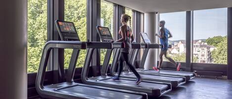 A gym with treadmills aligned in a row, large windows, and a view of buildings and trees outside.