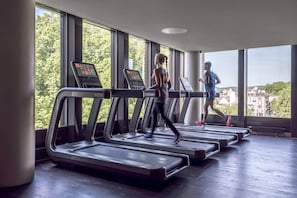 A gym with treadmills aligned in a row, large windows, and a view of buildings and trees outside.