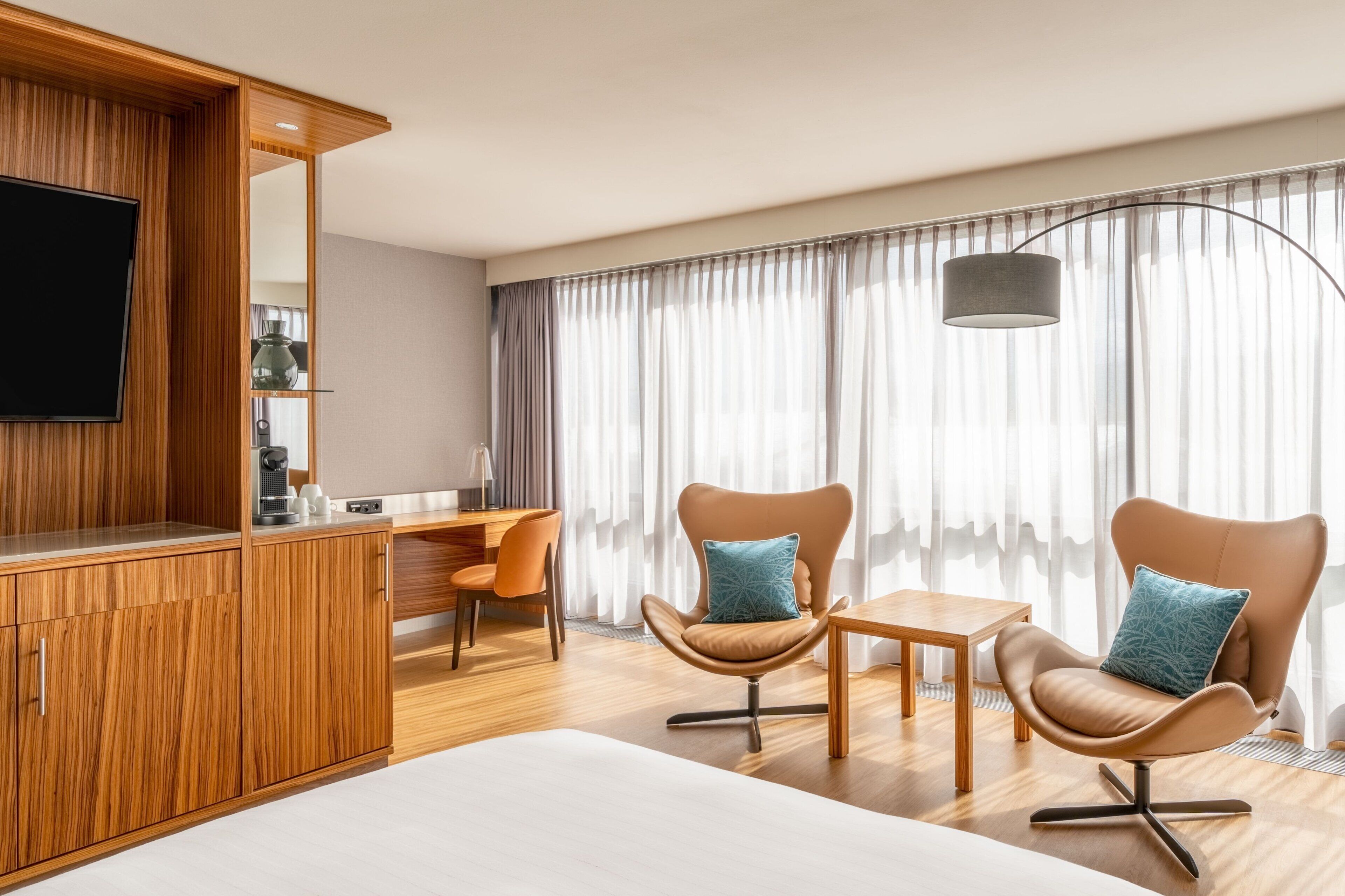A modern hotel room with a wooden desk, two armchairs, a TV, and a large window with curtains.