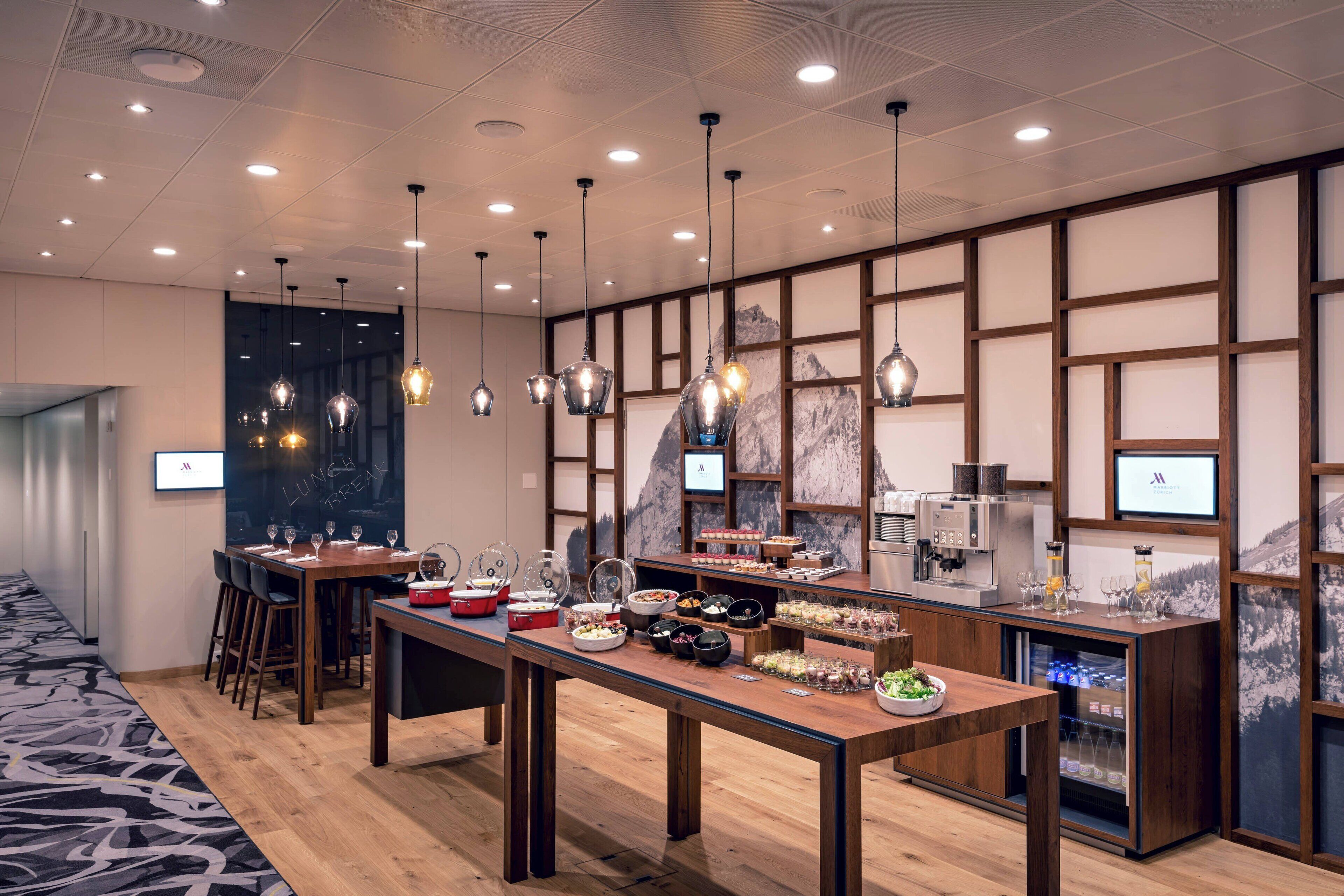 A modern hotel dining area with wooden tables, hanging pendant lights, and a buffet setup.