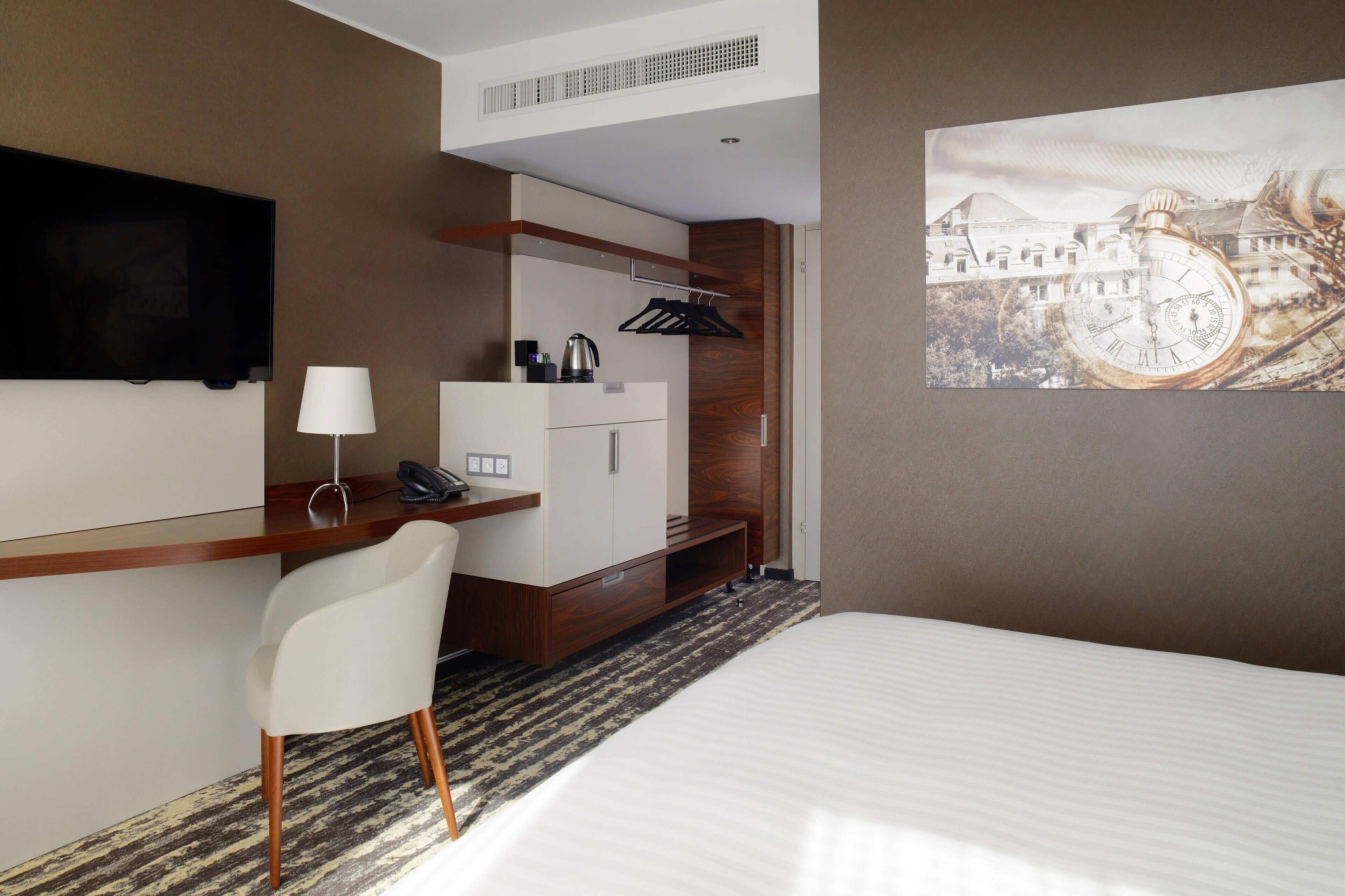 A hotel room with a bed, a desk with a chair, a TV, a coffee maker, and a painting on the wall.