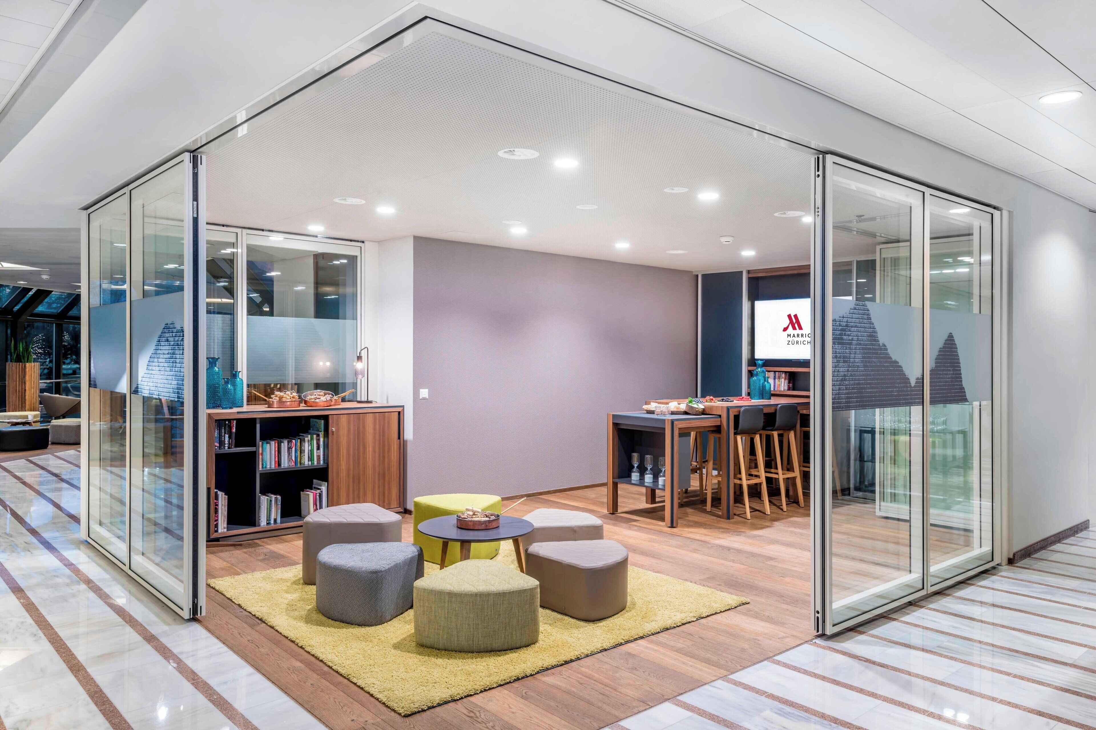 A modern office space with glass walls, a wooden bookshelf, a round table with chairs, and a small seating area with ottomans.