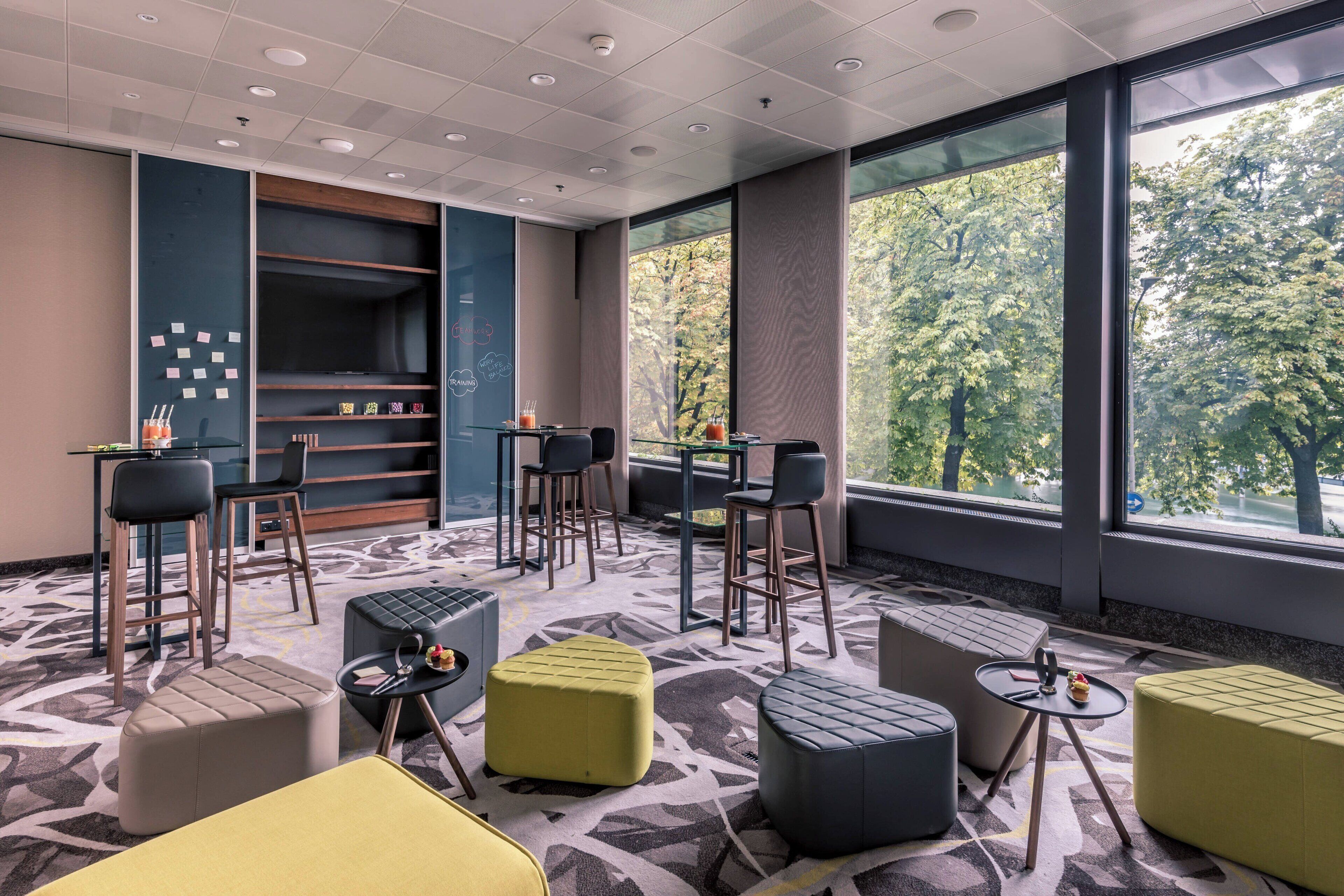 A modern lounge area with seating arrangements, a coffee bar, and large windows offering a view of trees.