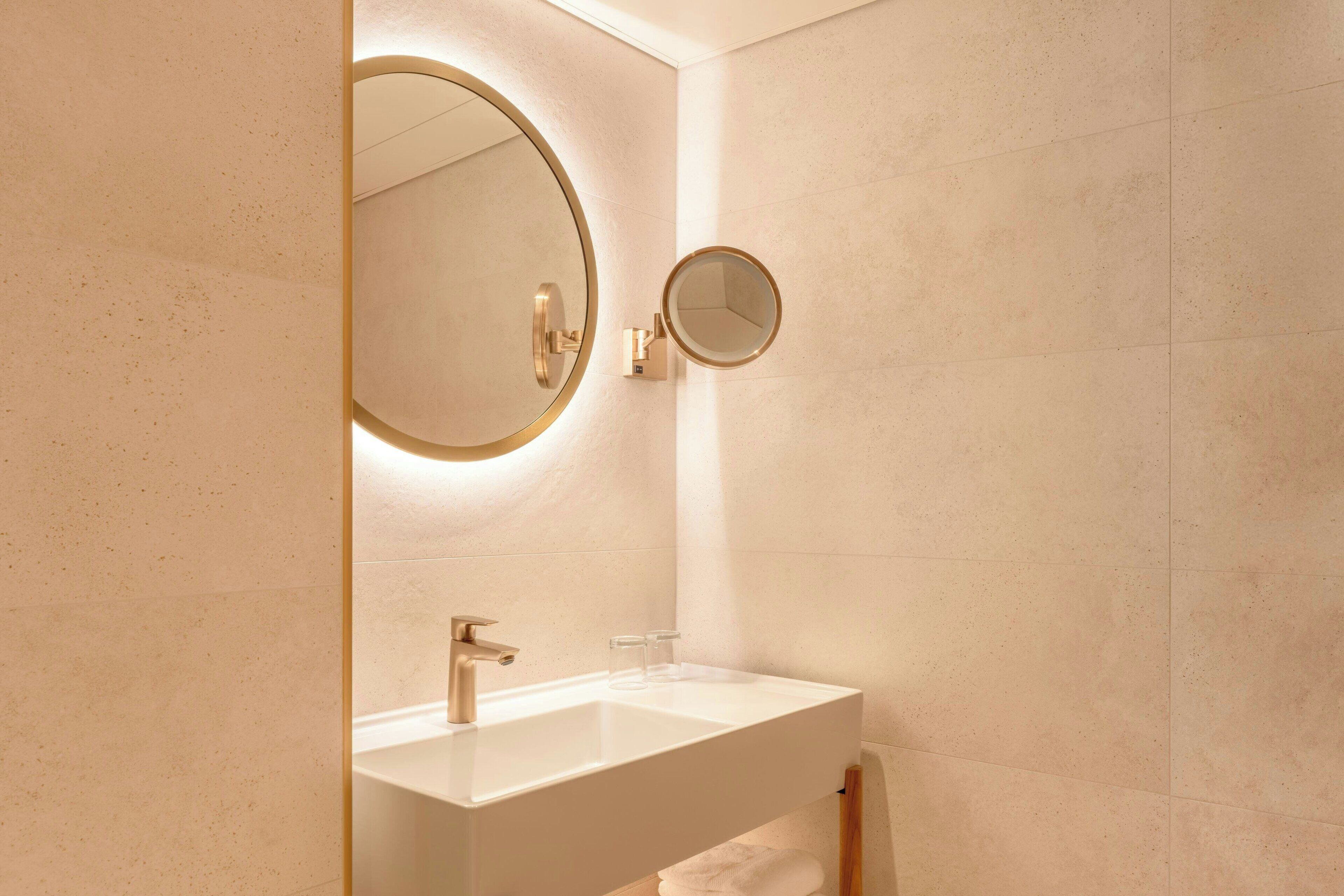 Une salle de bain avec un lavabo blanc, un robinet doré et un miroir rond doté d’un cadre doré.