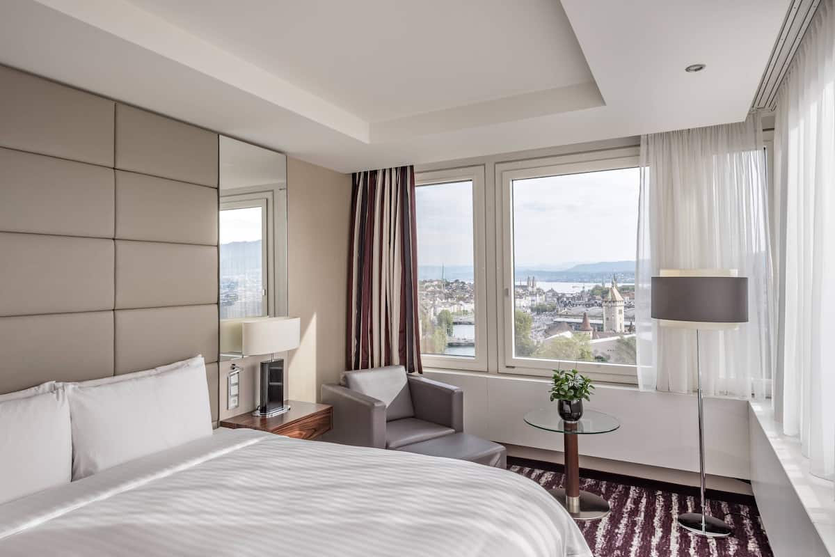 Hotellihuone, jossa on suuri sänky, oleskelualue tuolilla ja pöydällä, lamppu sekä näkymä kaupunkimaisemaan ikkunoista.