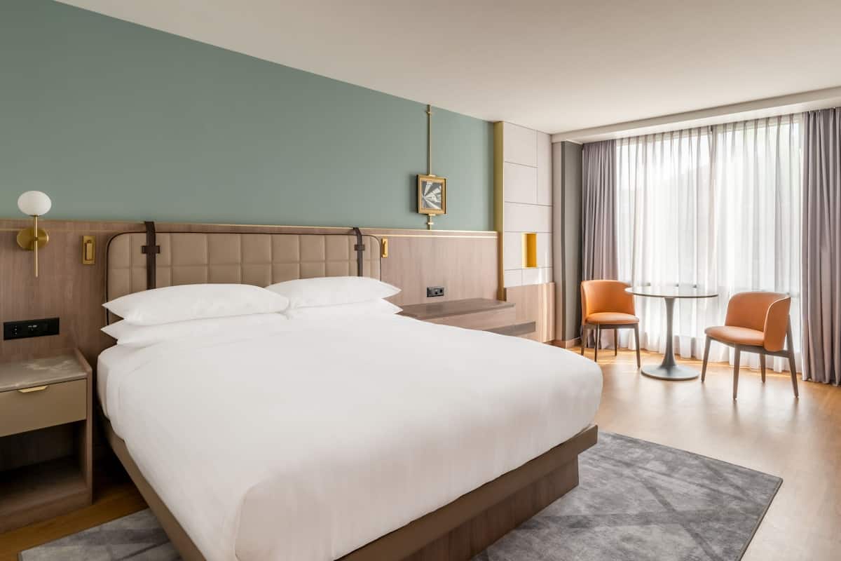 Hotellihuone, jossa on suuri sänky, yöpöydät, työpöytä, tuoli, pieni pöytä ja ikkuna, jossa on verhot.