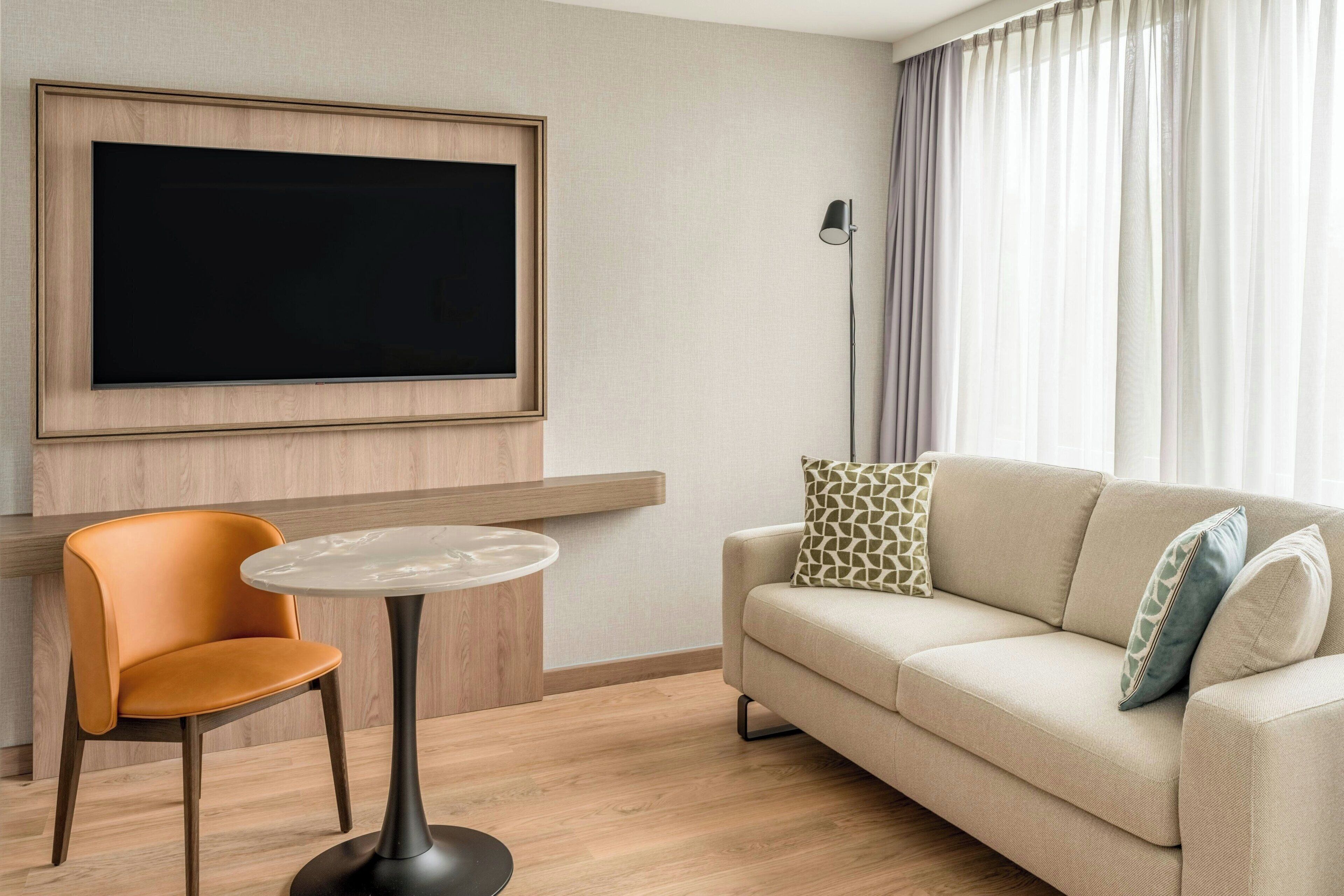 A modern living room with a flat-screen TV mounted on the wall, a round coffee table, a beige sofa with patterned cushions, and a wooden floor.