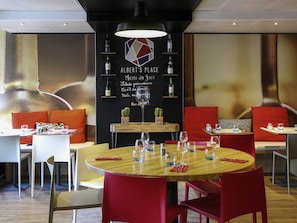 Interior de restaurante con mesas redondas, sillas rojas y una pared negra que muestra opciones de menú.