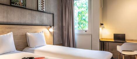 Habitación de hotel con dos camas, un escritorio con una computadora portátil, una silla, una ventana con cortinas y obras de arte colgadas en la pared.