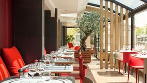 A restaurant with tables set for dining, red chairs, and a large tree in the center. - ibis Paris Gennevilliers (Gennevilliers)