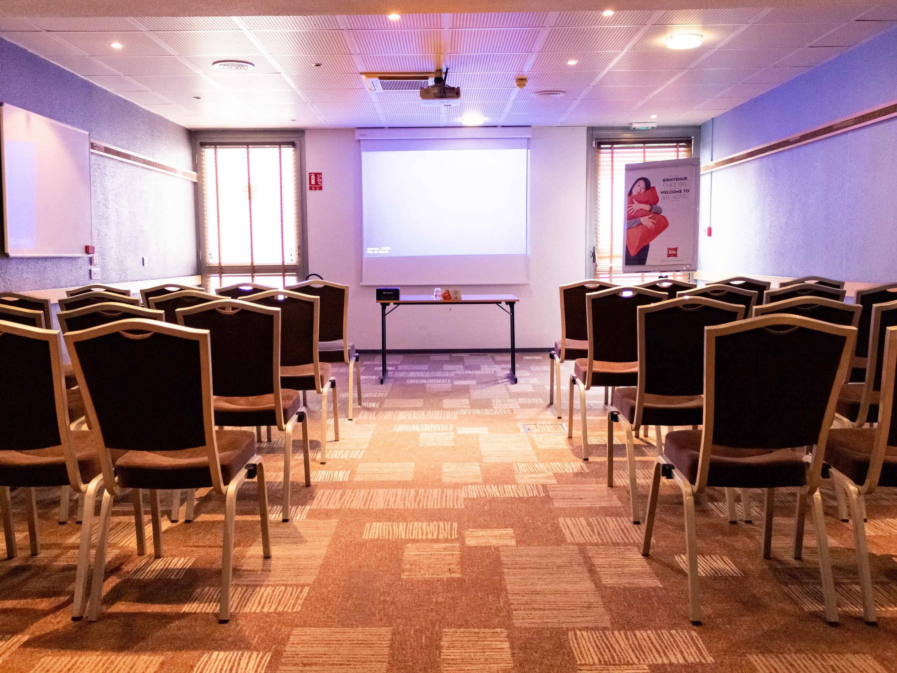 Une salle de conférence avec des rangées de chaises, un écran de projection et une table pour les présentations.