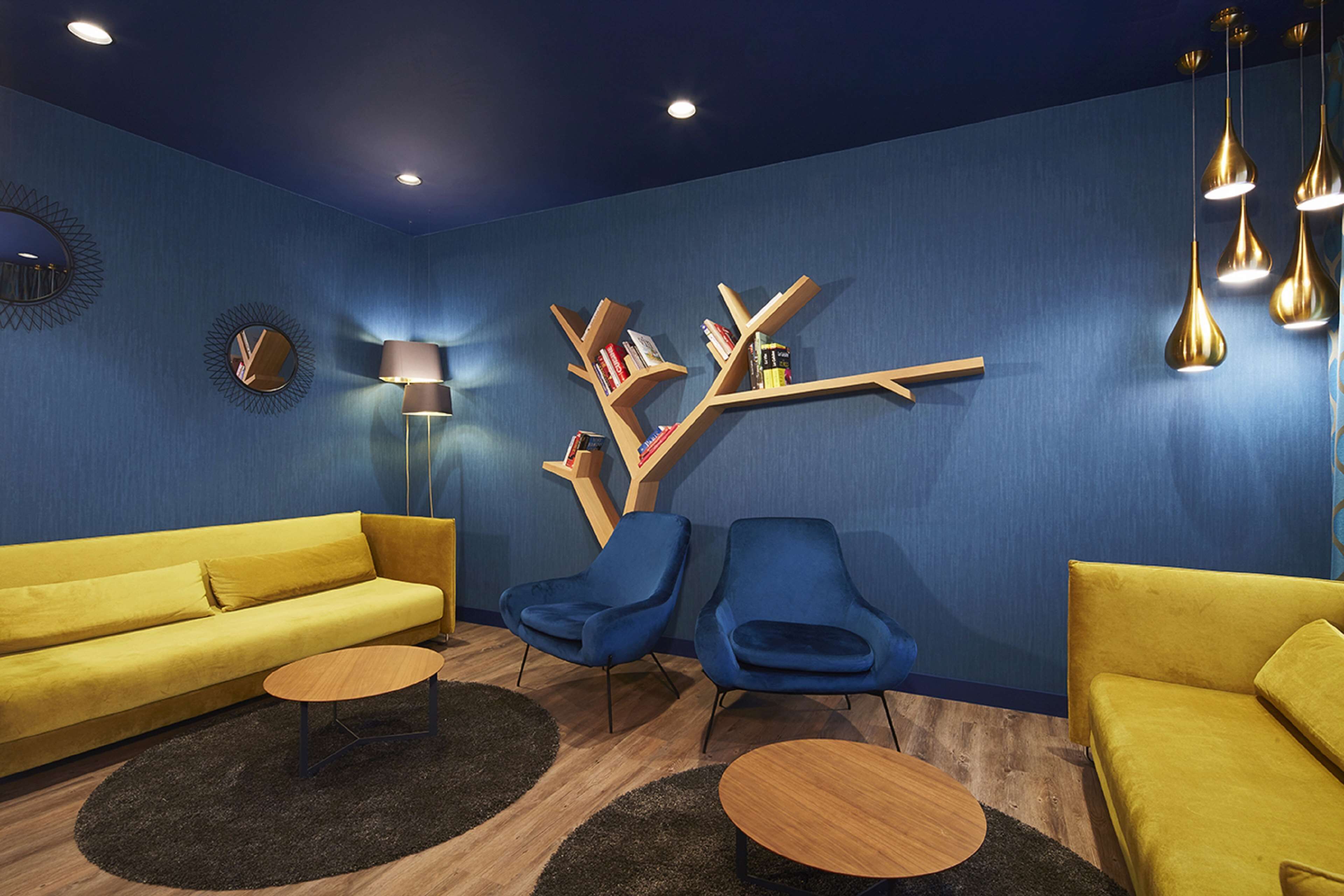 Una sala de estar moderna con pared azul, sofá amarillo, sillones azules y una estantería de madera.