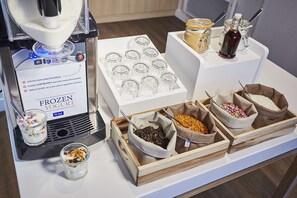A frozen yogurt station with a machine, cups, and various toppings in containers. - Hôtel Kyriad Paris Bezons - La Défense (Bezons)