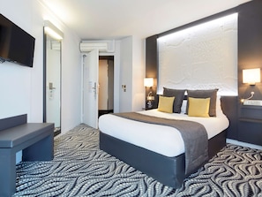 A modern hotel room with a large bed, a bedside table, a flat-screen TV mounted on the wall, and a patterned carpet. - Hôtel Kyriad Paris Bezons - La Défense (Bezons)