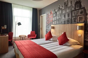 A hotel room with a large bed, red and white bedding, a desk with a chair, a small round table, and a chair. There is a window with curtains, a lamp, and a wall mural of buildings. - Huxley House Hotel (Amsterdam)
