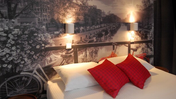 A hotel room with a bed, a bicycle, and a mural of a cityscape. - Huxley House Hotel (Amsterdam)