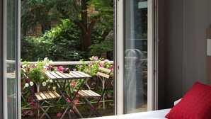 A room with a bed, a red pillow, and a view of a balcony with a wooden table and chairs, surrounded by flowers and greenery. - Huxley House Hotel (Amsterdam)