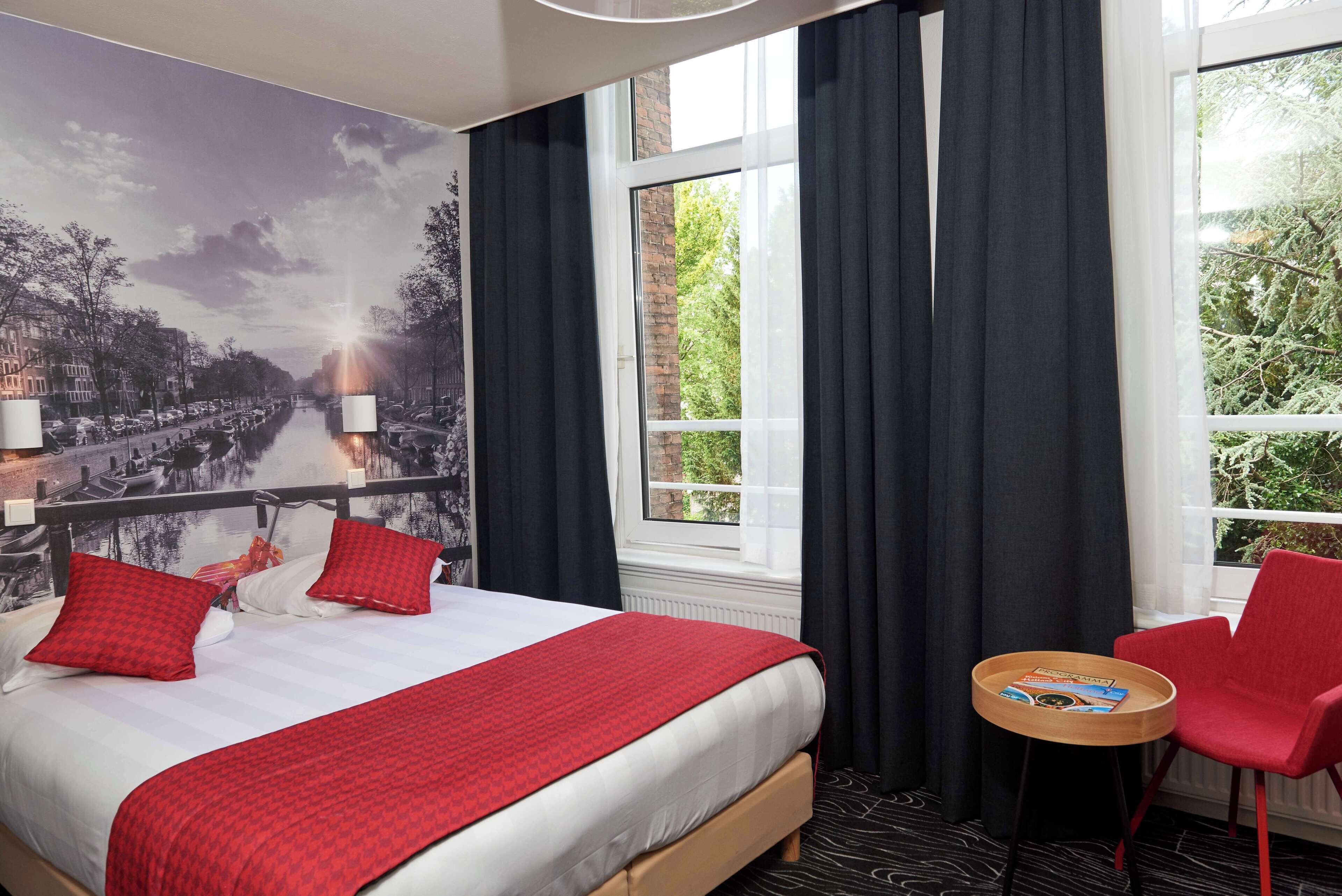 Een hotelkamer met een bed, een klein tafeltje met dienblad, een rode stoel en een groot raam met gordijnen.
