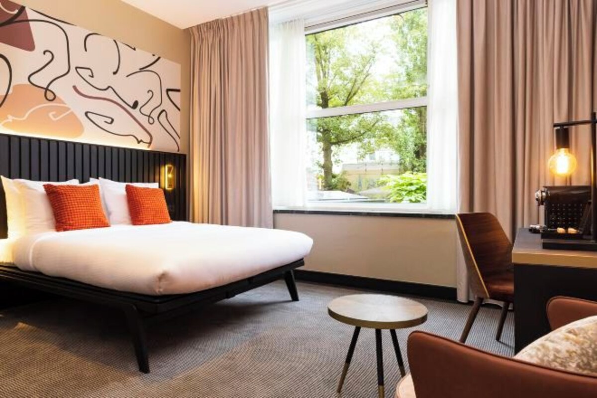 Ett modernt hotellrum med en stor säng, ett litet runt bord och utsikt över grönområden utanför.