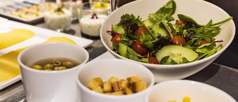Un bufé con diversos platos, incluyendo ensalada, aceitunas, maíz y un tazón que parece ser de ensalada de frutas.