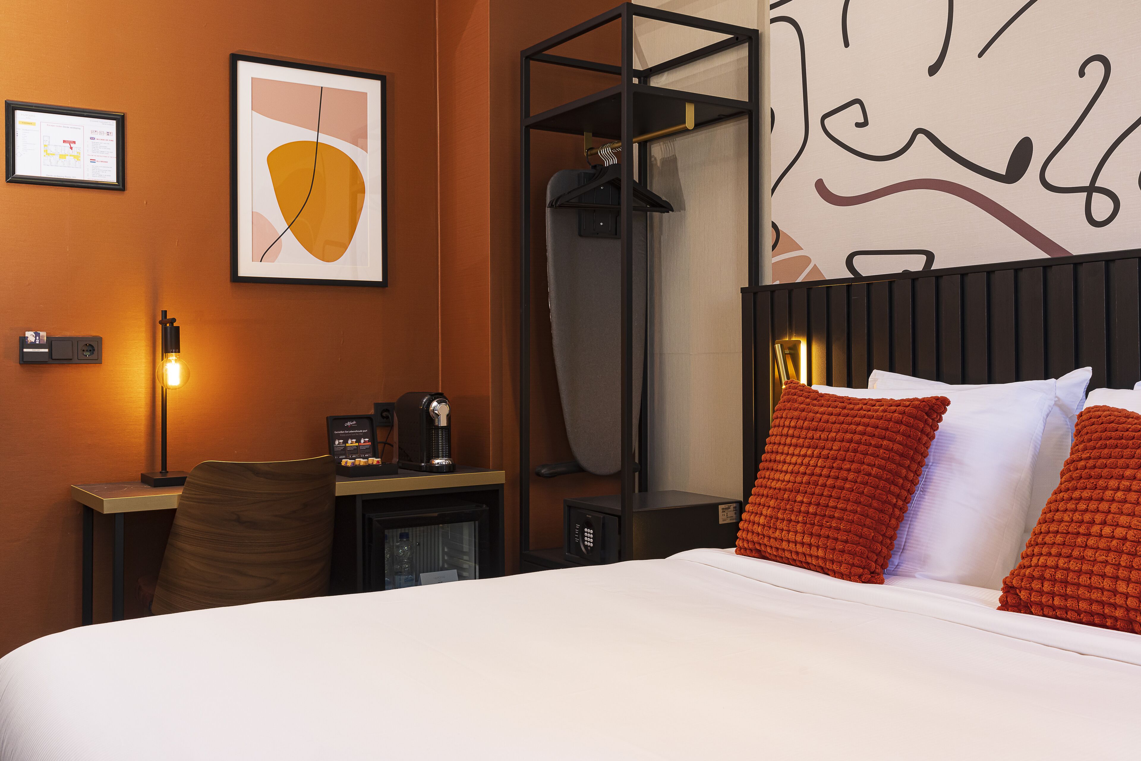 A hotel room with a bed, a desk with a coffee machine, a chair, a coat rack, and a framed picture on the wall.