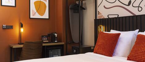 A hotel room with a bed, a desk with a coffee machine, a chair, a coat rack, and a framed picture on the wall.