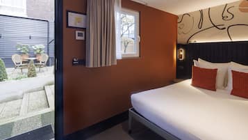 A hotel room with a bed, a window with curtains, a small table with chairs, and a view of the outside.