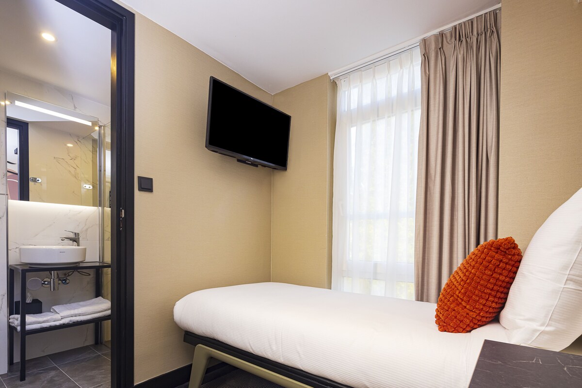 Habitación de hotel con cama, televisor, baño con lavamanos y ventana con cortinas.