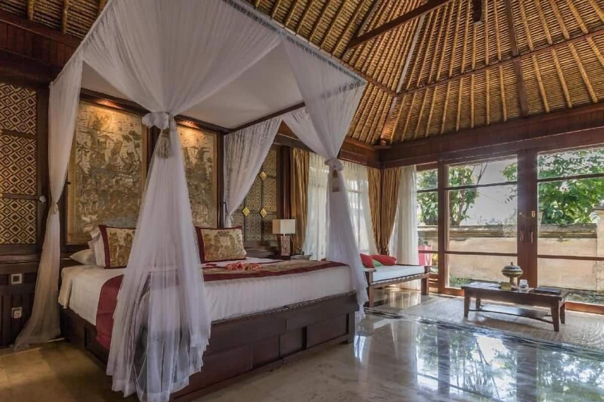 A four-poster bed with a canopy, a nightstand, a table with a lamp, and a view of greenery outside.