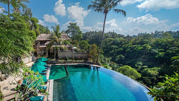 Una piscina infinita con borde de agua azul cristalina, rodeada de vegetación exuberante y un edificio con techo de paja.